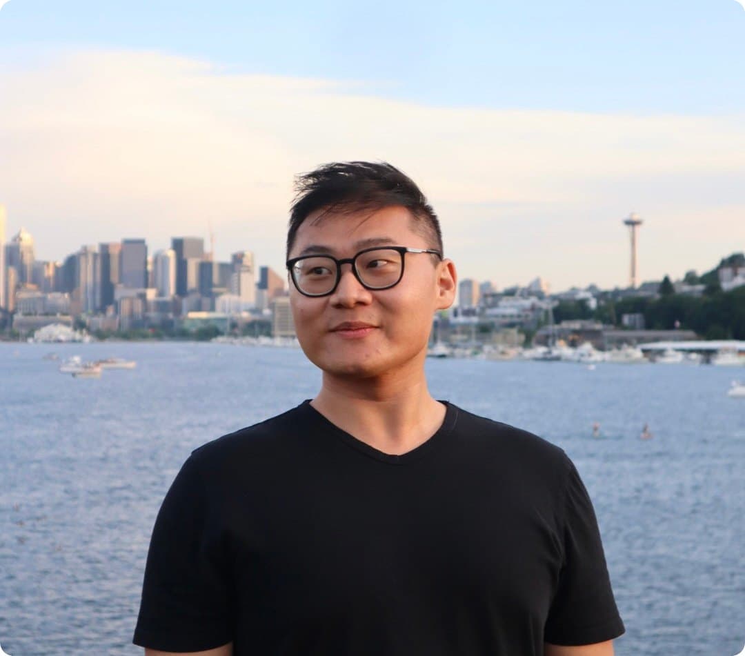 Andrew Ly smiling in a black t-shirt by the water with a city skyline behind him